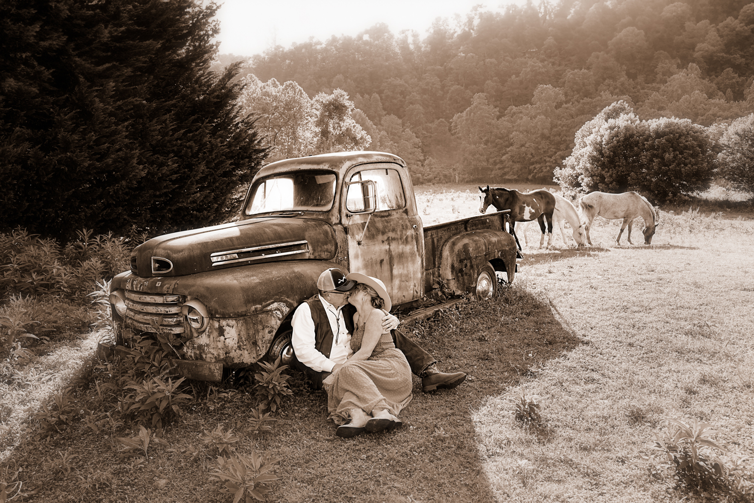 Sepia portrait of a couple sitting on the ground leaned against an old 1950 Ford truck kissing with horses in the background