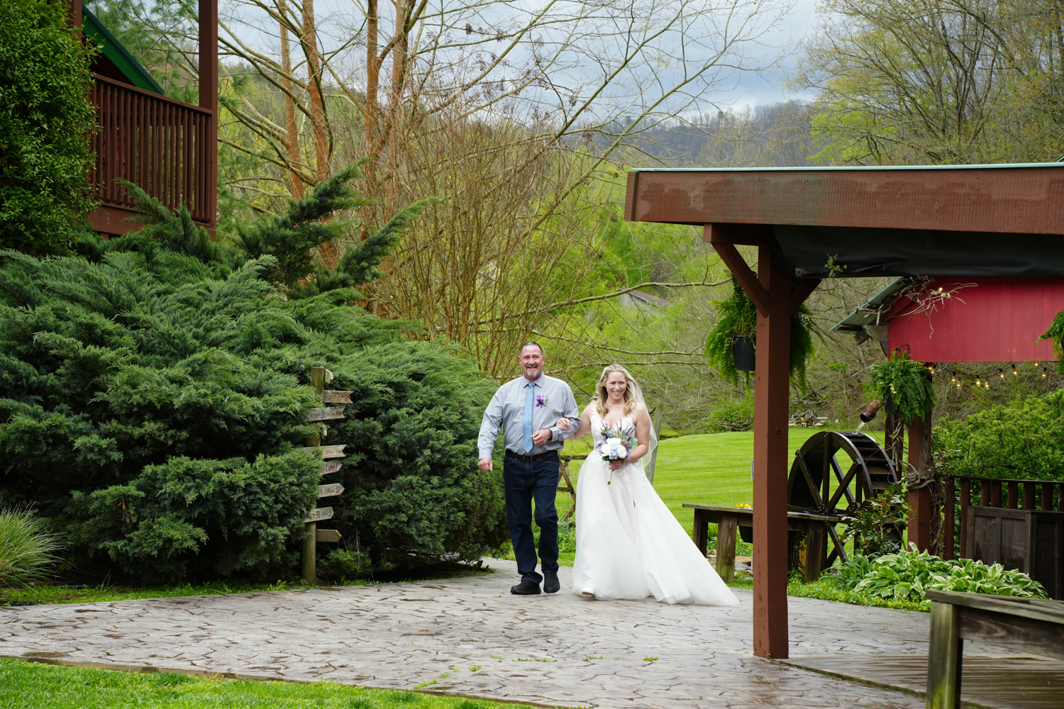 Father walking his daughter down the aisle on her wedding day along a stone sidewalk by a water wheel in the mountains of Pigeon Forge