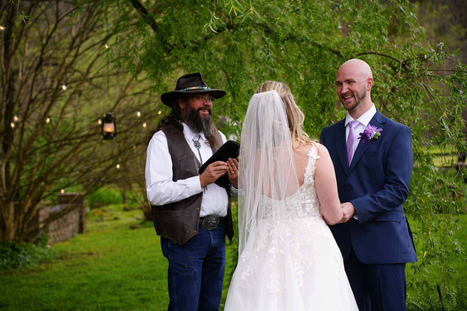 Preacher in a black hat and mountain vest performing a ceremony under a willow tree in early spring while the groom smiles at his bride