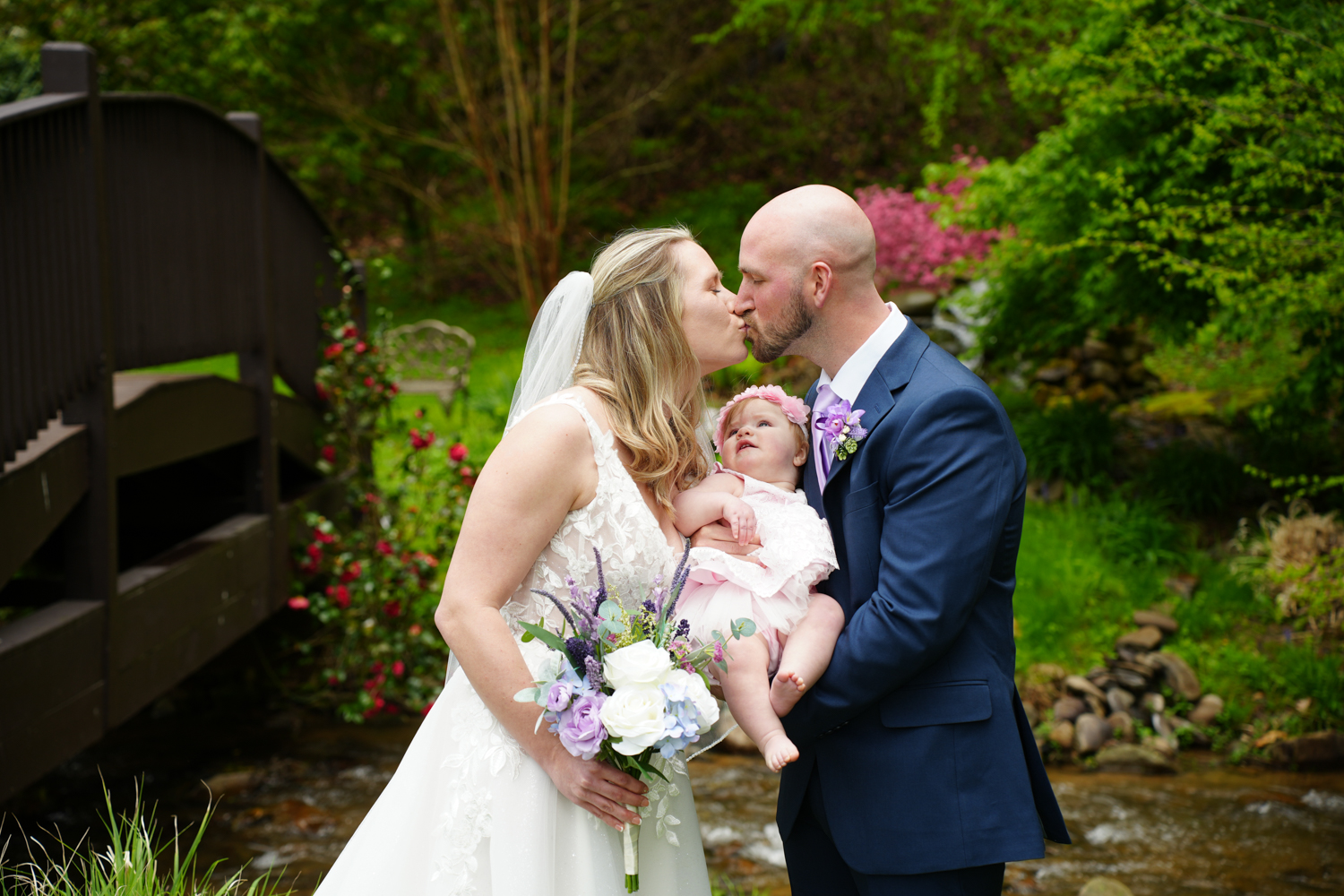 Bride and groom kissing while holding their baby girl who is looking up at them