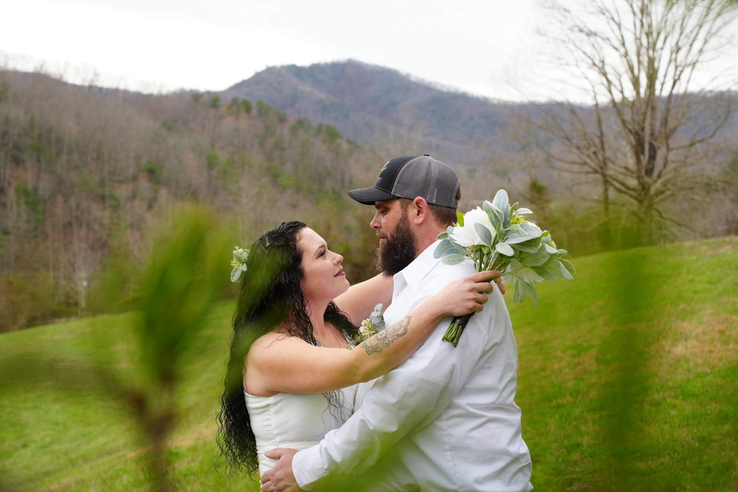 Groom in a baseball cap holding his bride behind evergreen branches in the mountains
