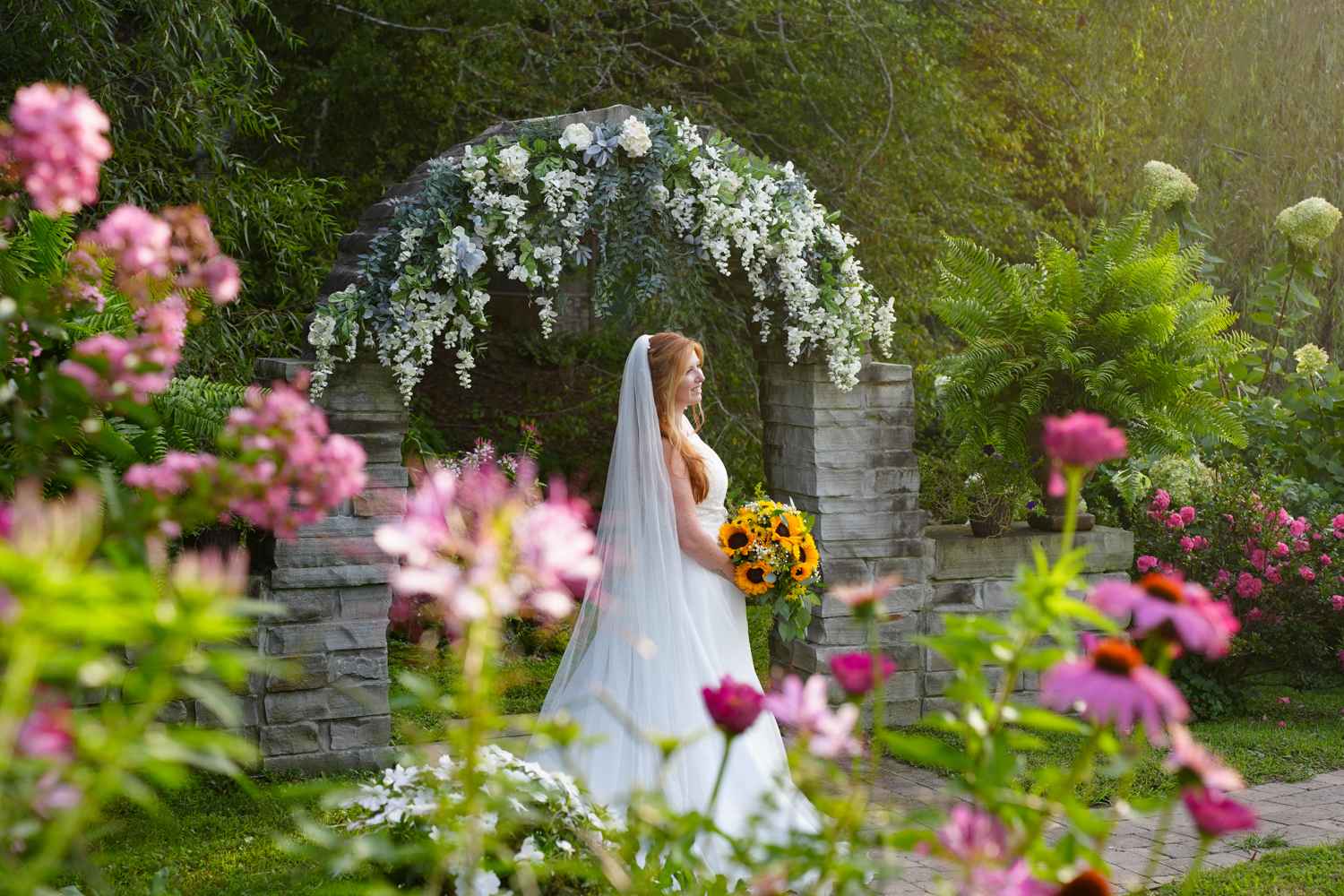 Bridal portrait at a stone arch with pink zinnia and other flowers in bloom