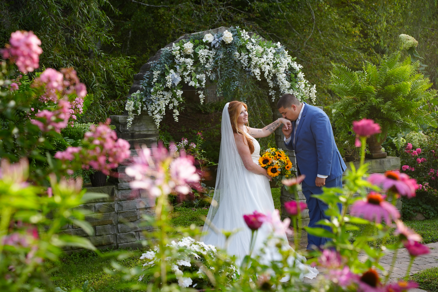 Pink flowers blooming at a stone arch where a groom is kissing his bride's hand
