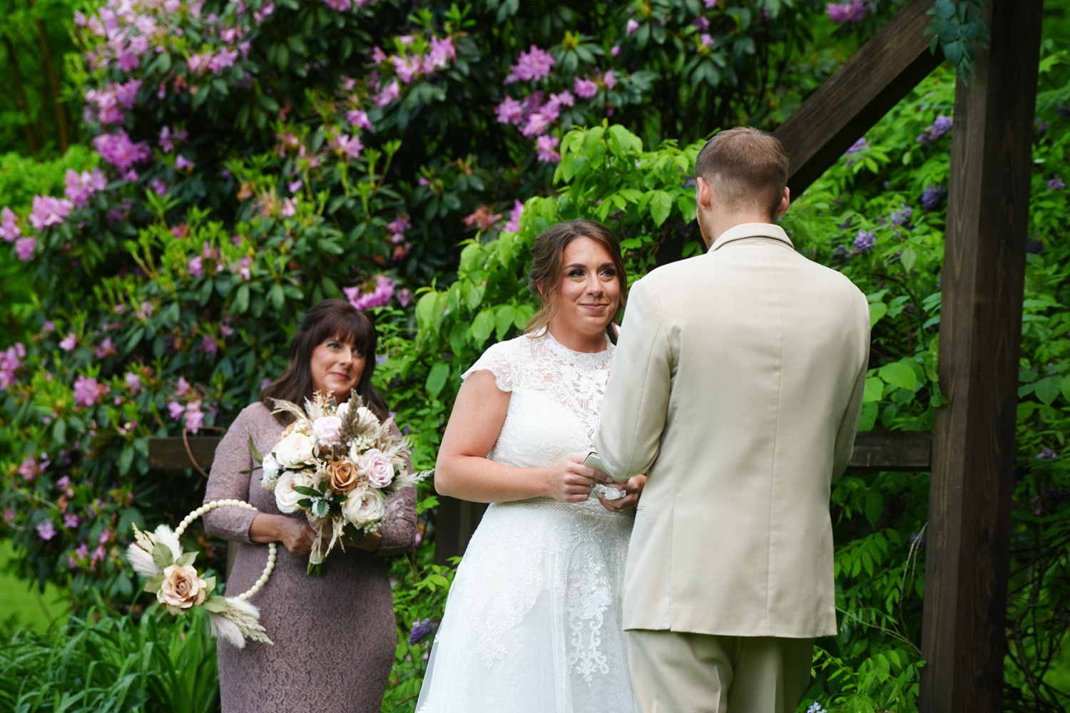 Bride saying her vows to her groom in the spring with purple rhododendrons in bloom and her mother watching lovingly from behind