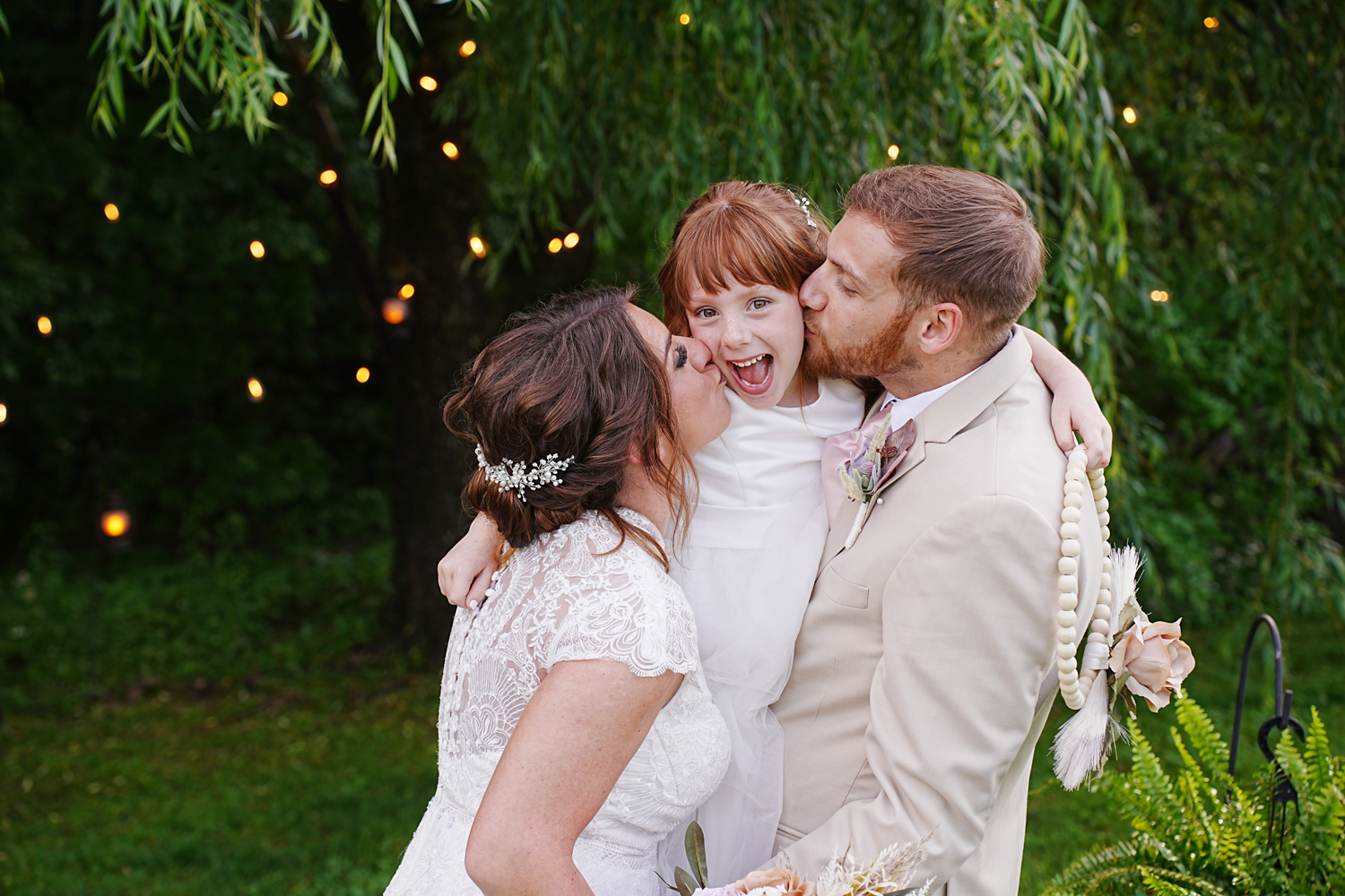 Wedding couple kissing their smiling happy daughter under a willow tree with twinkling lights