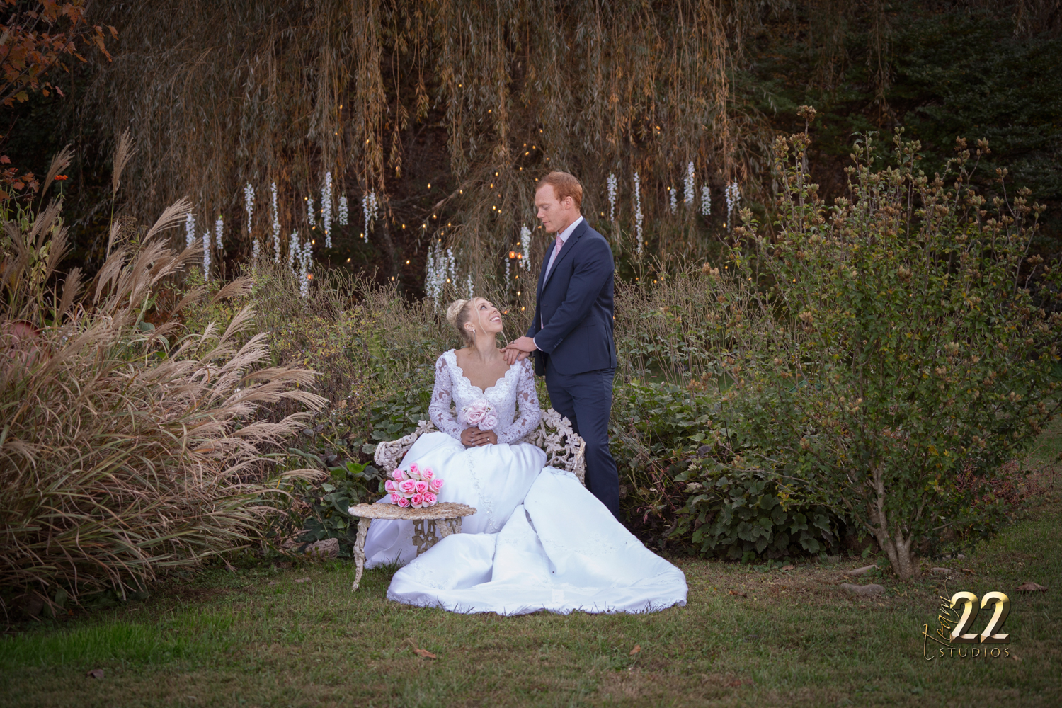 Bride seated looking up at her groom near a willow tree in the fall