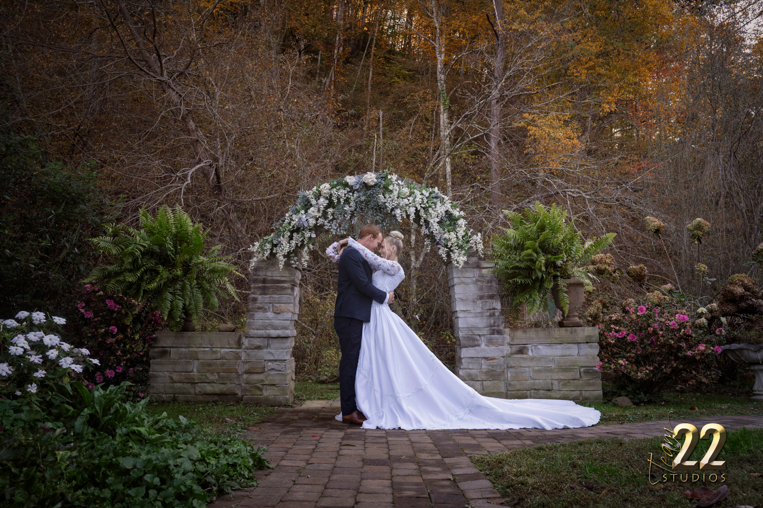 Bride with her arms around her groom at a stone archway with whie flowers in the fall