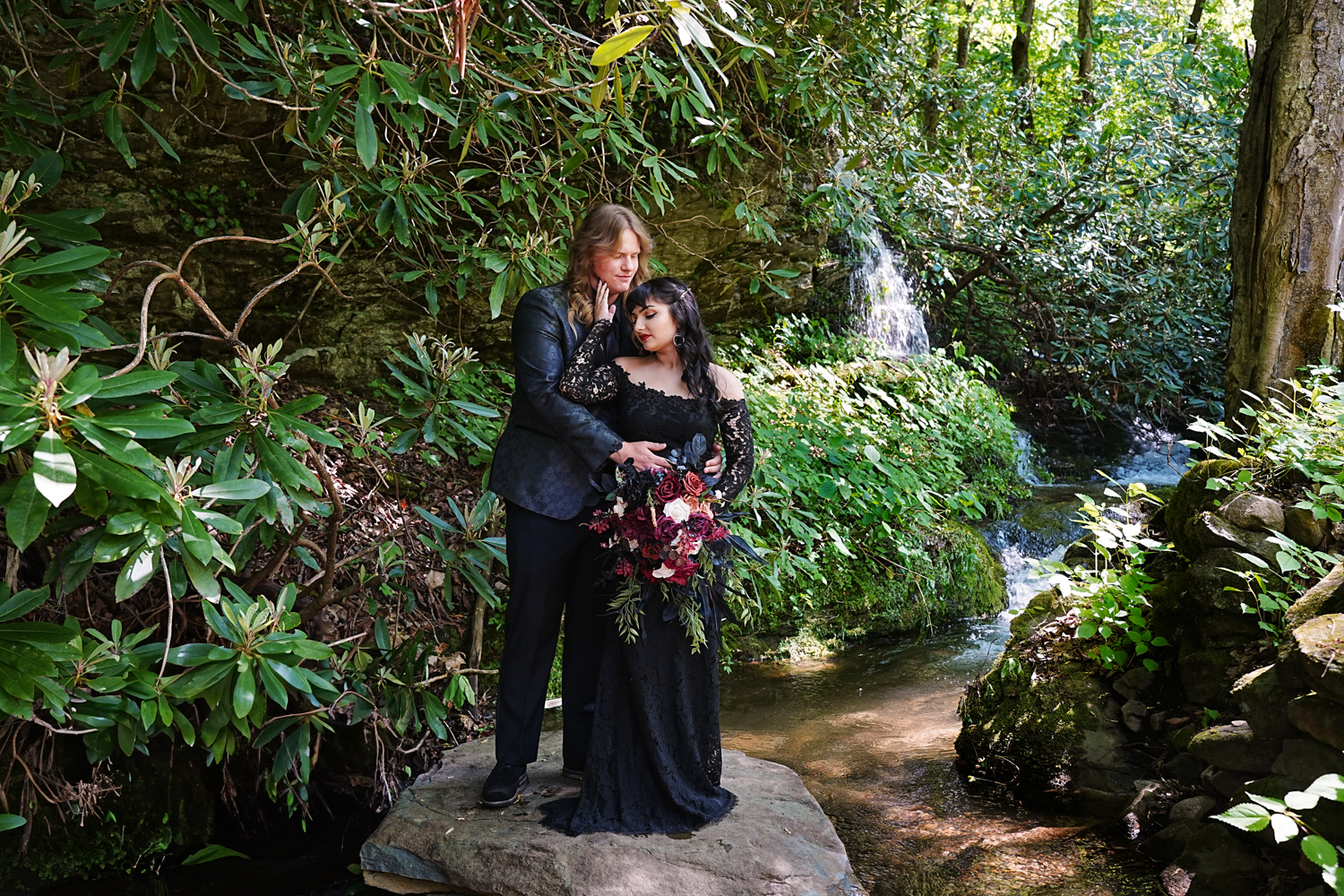 Bride in a black dress posing with her groom on a rock in the mountains by a waterfall