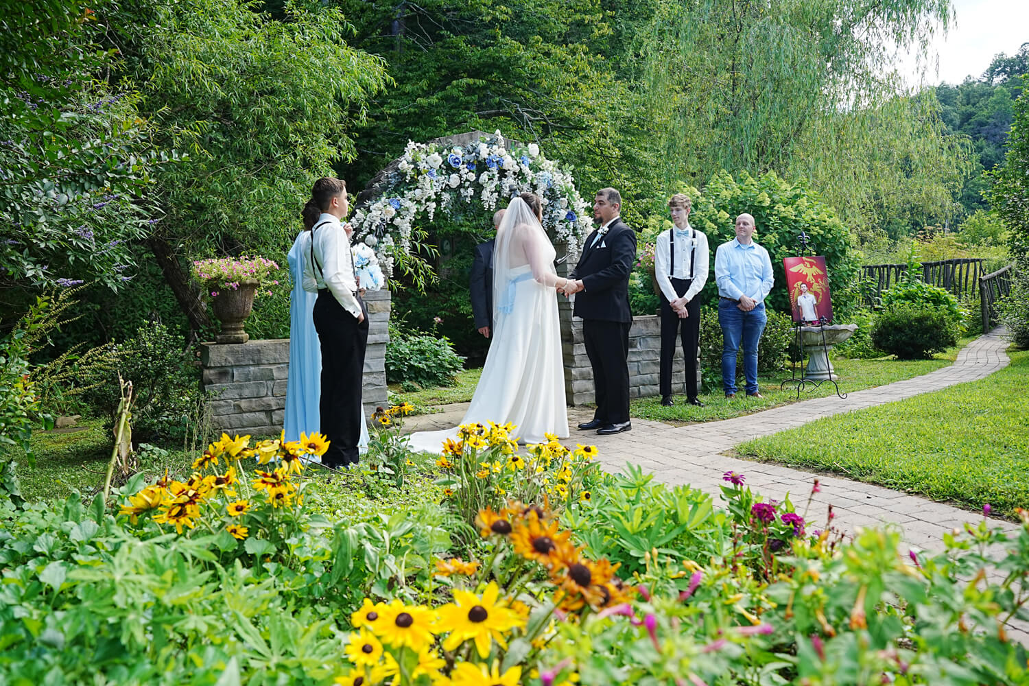 Wedding in the summer at a stone archway decorated in vines and flowers in front of a garden of flowers