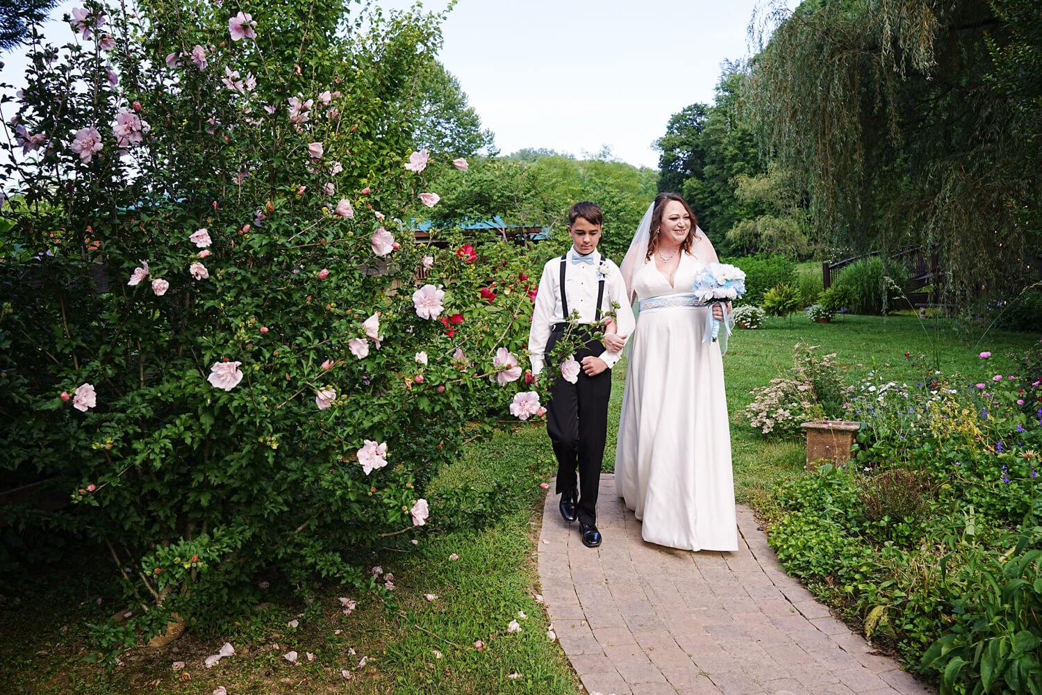 Son walking his mother down the aisle near a pink flowering rose of sharon bush in the summer