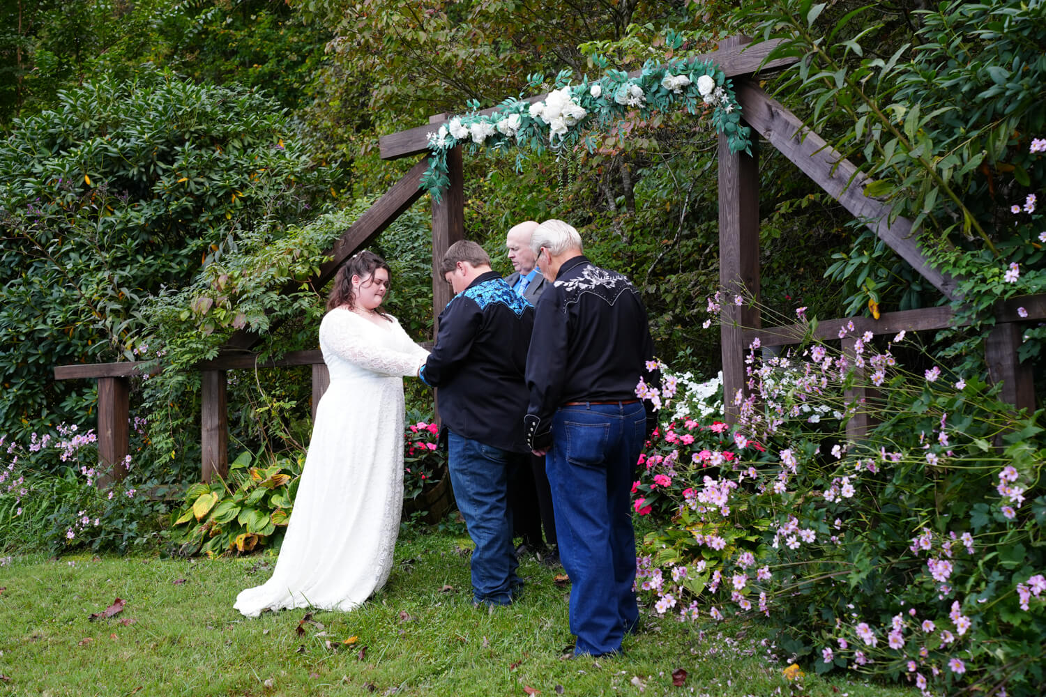 Wedding prayer at a wooden arbor with pink flowers in bloom all around them