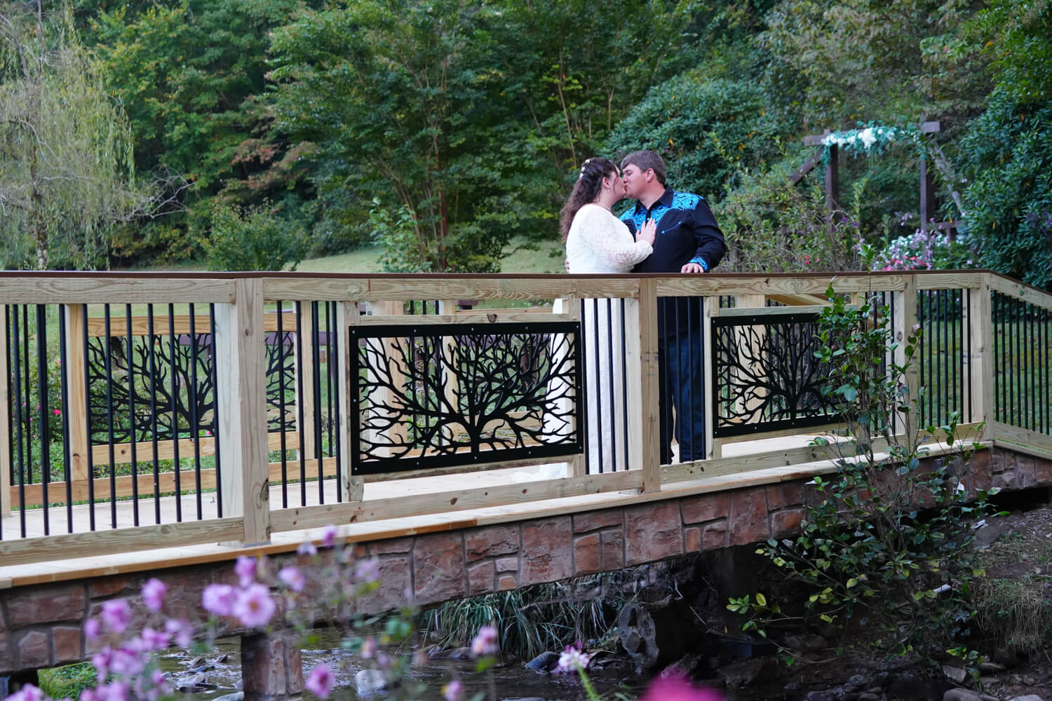 Wedding couple kissing on a formal bridge with stonework and iron tree panels above a creek with pink flowers growing next to it