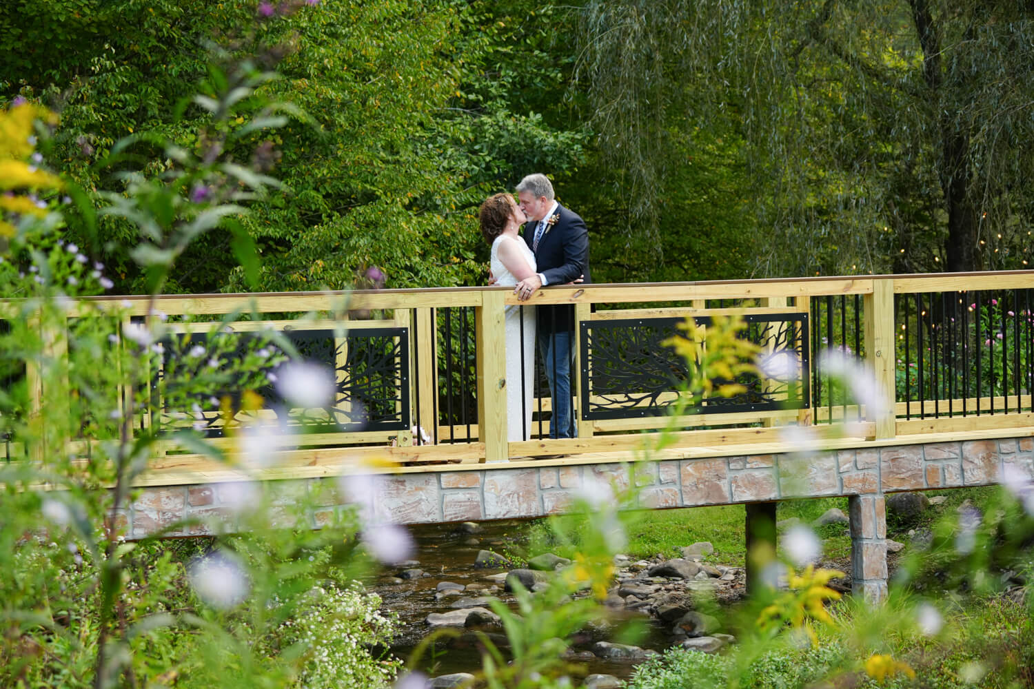 Formal wooden bridge with black iron willow tree panels and composite stone across a creek in the fall with a wedding couple kissing