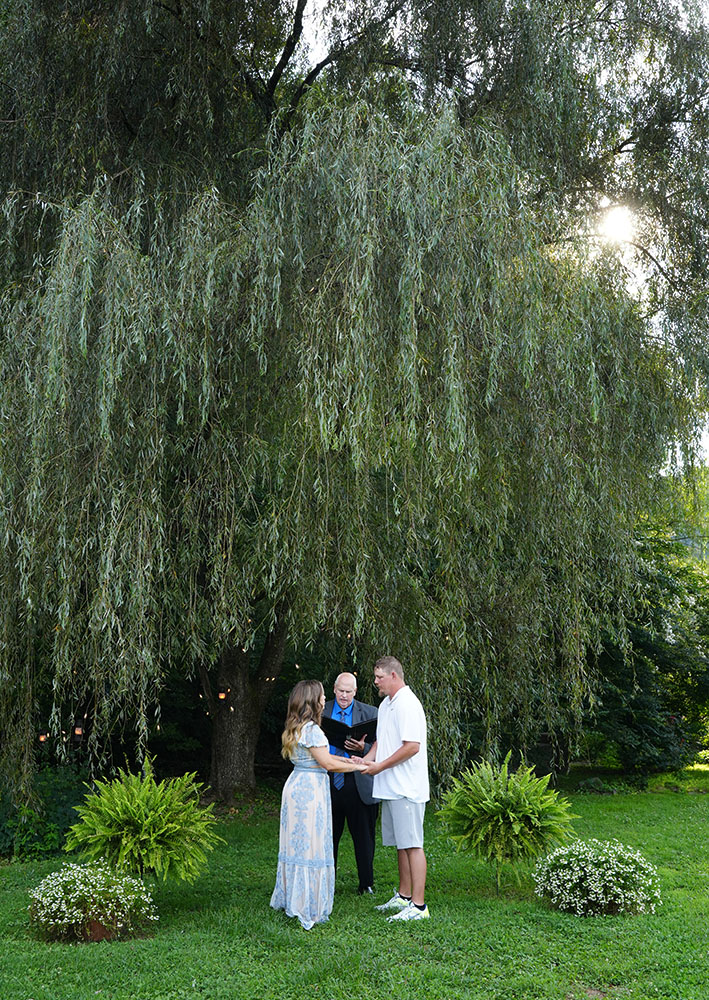 Couple in casual attire getting married under a willow tree in the summer with ferns and white flowers next to them