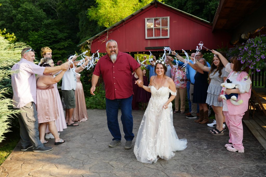 Couple exiting their wedding with guests waving streamers in front of a red cottage at the Honeysuckle Hills wedding venue