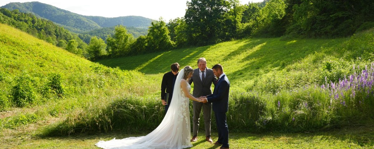Father leading prayer for his daughter and her groom in a wildflower meadow with purple larkspur in bloom with a mountain view behind them at the Honeysuckle Hills wedding venue in Pigeon Forge, TN