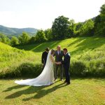 Father leading prayer for his daughter and her groom in a wildflower meadow with purple larkspur in bloom with a mountain view behind them at the Honeysuckle Hills wedding venue in Pigeon Forge, TN