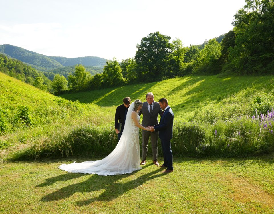 Father leading prayer for his daughter and her groom in a wildflower meadow with purple larkspur in bloom with a mountain view behind them at the Honeysuckle Hills wedding venue in Pigeon Forge, TN