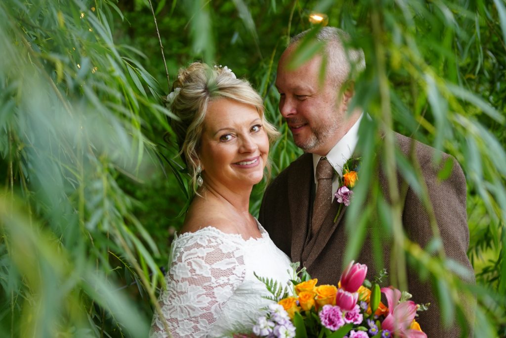 Bride smiling through the romantic branches of a willow as her husband looks at her adoringly at Honeysuckle Hills